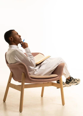 Gestures of diligence and excellence, reviewing lessons for studying and school tests, developing educational curricula in Saudi Arabia, a side portrait of a smiling Arab Gulf Saudi teenager wearing traditional attire, reclining on a chair holding a textbook and a pen, with a white background.