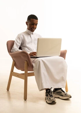 بورتريه لشاب عربي سعودي خليجي مبتسم يرتدي الثوب التقليدي يجلس على كرسي مريح و يمسك بيده اللابتوب، تعابير تدل على الاستمتاع باستخدام الحاسب المحمول، دردشة وتواصل عبر الانترنت, استخدام الاجهزة والتقنيات الحديثة والمتطورة,، خلفية بيضاء