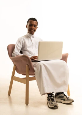 تعابير تدل على الاستمتاع باستخدام الحاسب المحمول، دردشة وتواصل عبر الانترنت, استخدام الاجهزة والتقنيات الحديثة والمتطورة, بورتريه لشاب عربي سعودي خليجي مبتسم يرتدي الثوب التقليدي يجلس على كرسي مريح و يمسك بيده اللابتوب، خلفية بيضاء