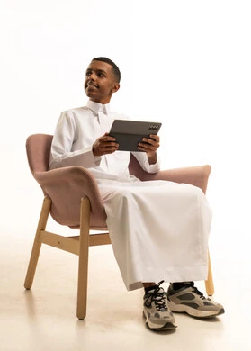 التواصل مع الاهل والاصدقاء، استخدام الأجهزة والتقنيات الحديثة، بورتريه لشاب عربي خليجي سعودي يرتدي الثوب التقليدي يجلس على كرسي مريح يمسك التابلت بيده بإيماءات السرور، تصفح مواقع التواصل الاجتماعي، خلفية بيضاء