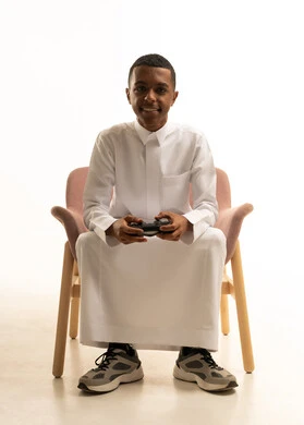 Interaction and immersion with PlayStation games, an enjoyable and exciting entertainment experience on modern devices, a portrait of a smiling young Arab Gulf Saudi man wearing traditional attire sitting on a chair holding a remote control in his hand, white background.