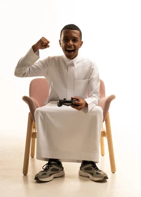 تعابيرالاستمتاع و الفوز بالألعاب الالكترونية الترفيهية، بورتريه لشاب عربي خليجي سعودي مبتسم يرتدي الثوب التقليدي يجلس على كرسى يمسك بيده عصا التحكم عن بعد، خلفية بيضاء