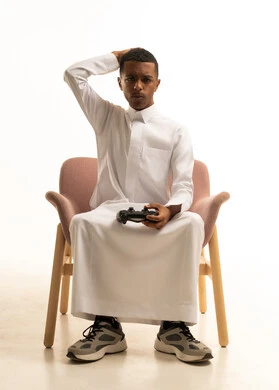 The use of modern technological devices, expressions indicating loss and defeat, a portrait of a young Arab Gulf Saudi man wearing traditional attire sitting on a chair holding a remote control in his hand, white background.