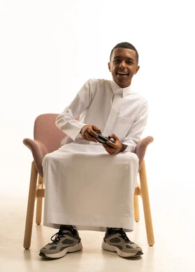 تجربة ترفيهية ممتعة ومثيرة في الأجهزة الحديثة، بورتريه لشاب عربي خليجي سعودي مبتسم يرتدي الثوب التقليدييجلس على كرسى يمسك بيده عصا التحكم عن بعد، خلفية بيضاء