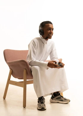 إستخدام أجهزة تقنية حديثة، بورتريه لشاب عربي خليجي سعودي مبتسم يرتدي الثوب التقليدي يضع سماعة الرأس يمسك بيدها عصا التحكم عن بعد، إيماءات وجه ويدين مختلفة تدل على الذهول والحماس، خلفية بيضاء