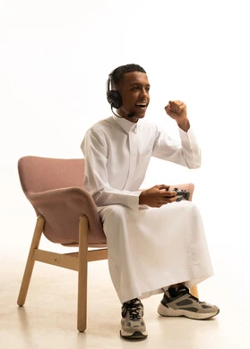 Facial expressions indicating victory and interaction with the game, using modern technological devices, a portrait of a smiling young Arab Gulf Saudi man wearing traditional attire, wearing headphones and holding a remote control in his hand, with a white background.