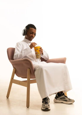 الإصغاء والاستمتاع باستخدام الهيدفون، قضاء أوقات ممتعة ومسلية، الاستماع إلى المواهب الغنائية الفنية، بورتريه لشاب عربي خليجي سعودي مبتسم يرتدي الثوب التقليدي يضع سماعة الرأس ويستمتع بتناول الفوشار، خلفية بيضاء