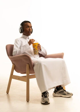 قضاء أوقات ممتعة ومسلية، الإصغاء والاستمتاع باستخدام الهيدفون، الاستماع إلى المواهب الغنائية الفنية، بورتريه لشاب عربي خليجي سعودي مبتسم يرتدي الثوب التقليدي يضع سماعة الرأس ويستمتع بتناول الفوشار، خلفية بيضاء