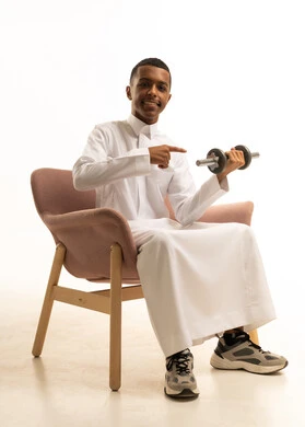 A portrait of a young Arab Gulf Saudi man wearing traditional attire sitting on a comfortable chair holding heavy weights in his hand, maintaining health, physical fitness, and agility, practicing sports and weightlifting with dumbbells, white background.
