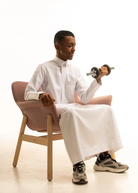 المحافظة على الصحة واللياقة الجسدية والرشاقة البدنية، بورتريه لشاب عربي خليجي سعودي يرتدي الثوب التقليدي يجلس على الكرسي المريح يحمل بيده الأوزان الحديدية الثقيلة، ممارسة التمارين الرياضية ورفع الأثقال مع الدمبل، خلفية بيضاء