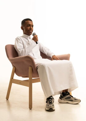 مواهب غنائية فنية، الاندماج مع الأغنية، تسجيل مقطع صوتي غنائي، شاب عربى خليجى سعودى يرتدى الثوب التقليدى يجلس على الكرسى يقوم بالغناء باستخدام الميكروفون بتعابير الحماس، خلفية بيضاء