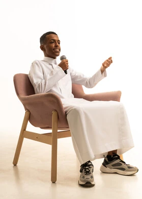 الاندماج مع الأغنية، تسجيل مقطع صوتي غنائي، مواهب غنائية فنية، شاب عربى خليجى سعودى يرتدى الثوب التقليدى يجلس على الكرسى يقوم بالغناء باستخدام الميكروفون بتعابير الحماس، خلفية بيضاء