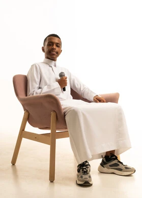 تسجيل مقطع صوتي غنائي، مواهب غنائية فنية، الاندماج مع الأغنية، شاب عربى خليجى سعودى يرتدى الثوب التقليدى يجلس على الكرسى يقوم بالغناء باستخدام الميكروفون بتعابير الحماس، خلفية بيضاء