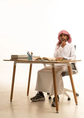 التركيز في حل وكتابة الواجب، التربية والتعليم بالمناهج المقررة للطلاب في المملكة العربية السعودية،  بورتريه لطالب عربي خليجي سعودي يرتدي الثوب الابيض والشماغ يجلس على الطاولة يقوم بالمذاكرة, خلفية بيضاء