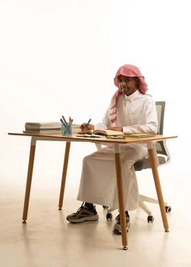 تأدية الواجبات المدرسية، التعليم الدراسي في السعودية، العودة إلى المدارس، بورتريه لطالب عربي خليجي سعودي يرتدي الثوب التقليدي والشماغ يجلس على الطاولة يقوم بتدوين الدروس ، خلفية بيضاء