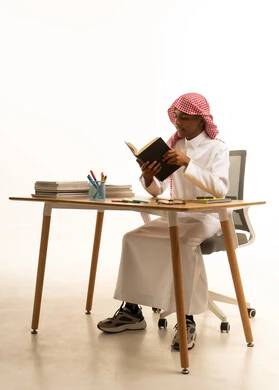 A portrait of a Saudi Gulf Arab student wearing traditional attire and a shemagh sitting at a table holding a school book, representing the concepts of learning, excellence, and diligence, the curricula and educational materials in Saudi Arabia, with a white background.