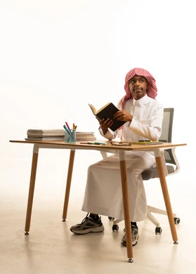 محاولة تذكر شيء ما، بورتريه لطالب عربي خليجي سعودي يرتدي الثوب التقليدي والشماغ يجلس على الطاولة يحمل كتاب مدرسى، المناهج والمواد التعليمية فى السعودية، خلفية بيضاء