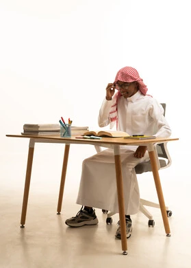 Gestures of diligence, focus, and love for learning, reviewing daily lessons, a portrait of a Saudi Gulf Arab student wearing traditional attire, a ghutrah, and glasses sitting at a table, with a white background.