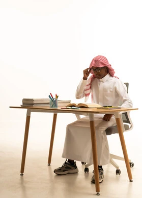 Daily lesson review, gestures of diligence, focus, and love for learning, a portrait of a Gulf Arab Saudi student wearing traditional attire, a ghutrah, and glasses sitting at a table, with a white background.
