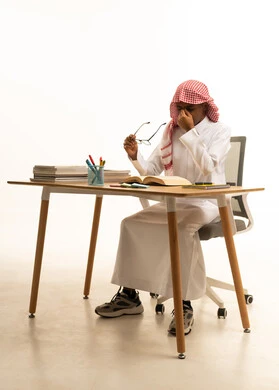 Diligence, focus, and a love for learning, a portrait of a Gulf Arab Saudi student wearing traditional attire and a shemagh sitting at a table taking a break while studying, a collection of pens and school books, education in the Kingdom of Saudi Arabia, white background.
