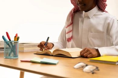المقررات والمناهج التعليمية الحديثة والمطورة بالسعودية، بورتريه مقرب ليد طالب عربي خليجي سعودي يرتدي الثوب التقليدي والشماغ يجلس على الطاولة يقوم بتدوين الملاحظات، خلفية بيضاء