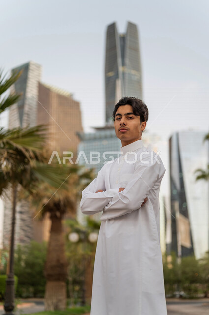 شاب عربي خليجي سعودي بإيماءات و تعابير وجه و يدين مختلفة يقف أمام أبراج مدينة الملك عبدالله المالية ( كافد ) في مدينة الرياض في المملكة العربية السعوية  ، إطلالة رائعة لأبراج المدينة ، جولة سياحية في المدينة
