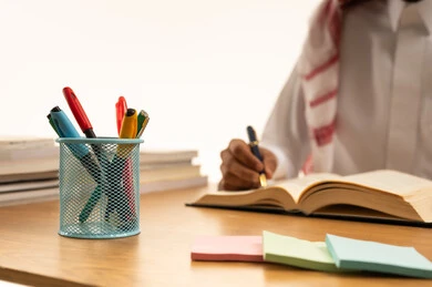 بورتريه مقرب ليد طالب عربي خليجي سعودي يرتدى الثوب والشماغ يجلس على الطاولة، مراجعة الدروس اليومية، مجموعة من الأقلام والكتب المدرسية، التعليم فى المملكة العربية السعودية، خلفية بيضاء