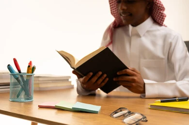 بورتريه مقرب لطالب عربي خليجي سعودي يرتدى الثوب والشماغ يجلس على الطاولة يقوم بقراءة كتاب، مجموعة من الأقلام والكتب المدرسية، التعليم فى المملكة العربية السعودية، تحضيرالدروس للوصول إلى النجاح، خلفية بيضاء