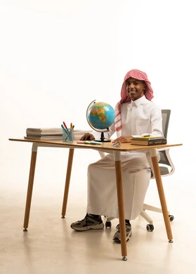 Education in the Kingdom of Saudi Arabia, a portrait of an Arab Gulf Saudi student wearing traditional attire and a shemagh sitting at a table looking at a globe model, the concept of research and exploration around the world, white background.