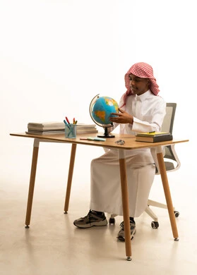 Using tools and aids, a portrait of a Saudi Gulf Arab student wearing traditional attire and a ghutrah sitting at a table holding a globe model, a collection of pens and school books, education in the Kingdom of Saudi Arabia, white background.