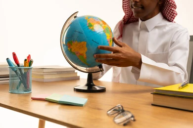 The concept of homeschooling, a portrait of an Arab Gulf Saudi student wearing traditional attire and a shemagh sitting at a table holding a globe model, a collection of pens and school books, education in the Kingdom of Saudi Arabia, white background.
