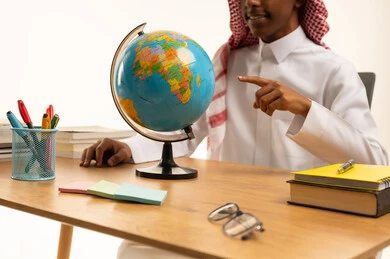 مفهوم البحث والإستكشاف حول العالم، بورتريه لطالب عربي خليجي سعودي يرتدي الثوب التقليدي والشماغ يجلس على الطاولة يشيرإلى مجسم الكرة الأرضية، مجموعة من الأقلام والكتب المدرسية، التعليم فى المملكة العربية السعودية، خلفية بيضاء