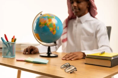 The importance of education in building a bright future for society, a portrait of a Saudi Arabian Gulf Arab student wearing traditional attire and a shemagh sitting at a table looking at a globe model, a collection of pens and school books, education in the Kingdom of Saudi Arabia, white background.