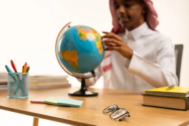 A portrait of a Saudi Gulf Arab student wearing traditional attire and a shemagh sitting at a table looking at a globe model, a collection of pens and school books, the concept of research and exploration around the world, academic education in Saudi Arabia, back to school, white background.