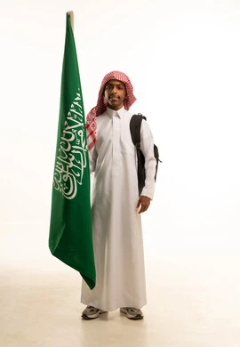 بورتريه لطالب عربي خليجي سعودي مبتسم يرتدي الثوب التقليدي والشماغ وحقيبة مدرسية يقف باستقامة، تلميذ مجتهد يحمل بيده علم المملكة العربية السعودية، إحياء ذكرى اليوم الوطني السعودي في المدارس، الانتماء والاعتزاز باليوم الوطني، خلفية بيضاء