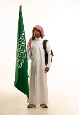 إحياء ذكرى اليوم الوطني السعودي في المدارس، الانتماء والاعتزاز باليوم الوطني، بورتريه لطالب عربي خليجي سعودي مبتسم يرتدي الثوب التقليدي والشماغ وحقيبة مدرسية، تلميذ مجتهد يحمل بيده علم المملكة العربية السعودية، خلفية بيضاء