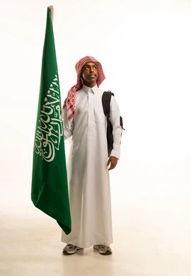 الانتماء والاعتزاز باليوم الوطني، احياء ذكرى اليوم الوطني السعودي في المدارس، بورتريه لطالب عربي خليجي سعودي مبتسم يرتدي الثوب التقليدي والشماغ وحقيبة مدرسية، تلميذ مجتهد يحمل بيده علم المملكة العربية السعودية، خلفية بيضاء