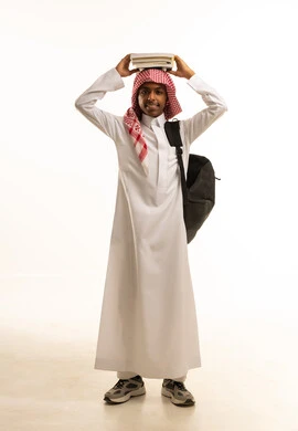 المدارس التعليمية في المملكة السعودية، بورتريه لفتى عربي خليجي سعودي مبتسم يرتدي الثوب التقليدي والشماغ وحقيبة ظهر يحمل فوق رأسه كتبه المدرسية, الاستعداد لبداية العام الدراسي الجديد، صورة الجسم بالطول الكامل، خلفية بيضاء