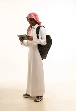 البحث عن معلومات مفيدة وهامة, بورتريه لشاب عربي خليجي سعودي مبتسم يرتدي الثوب التقليدي والشماغ وحقيبة ظهر يحمل بيده جهاز التابلت, دمج التقنية والتكنولوجيا مع التعلم، حضور دورات تعليمية الكترونية، صورة الجسم بالطول الكامل، خلفية بيضاء