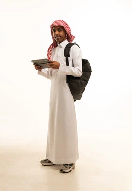 حضور دورات تعليمية الكترونية، بورتريه من الجانب لشاب عربي خليجي سعودي مبتسم يرتدي الثوب التقليدي والشماغ وحقيبة ظهر يحمل بيده جهازالتابلت, البحث عن معلومات مفيدة وهامة، النظر الى شىء ما، صورة الجسم بالطول الكامل، خلفية بيضاء
