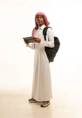 Integrating technology and tech with learning, a side portrait of a smiling young Arab Gulf Saudi man wearing traditional attire and a shemagh, carrying a backpack and holding a tablet in his hand, searching for useful and important information, looking at something, full-body image, white background.