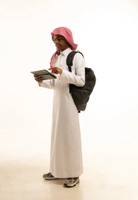 A side portrait of a smiling Saudi Gulf Arab student wearing traditional attire and a shemagh, carrying a backpack and holding a tablet in his hand, searching for useful and important information, integrating technology with learning, attending online educational courses, full-body image, white background.