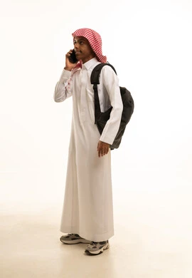 إجراء مكالمة هاتفية عبر الهاتف المحمول, بورتريه لطالب عربي خليجي سعودي مبتسم يرتدي الثوب التقليدي والشماغ وحقيبة ظهر يتحدث فى الجوال, خلفية بيضاء