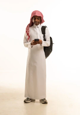 البحث والمطالعة من خلال الجوال، تصفح التطبيقات ووسائل التواصل الاجتماعي، استخدام الأجهزة والتقنيات الحديثة والمتطورة, بورتريه لشاب عربي خليجي سعودي مبتسم يرتدي الثوب التقليدي و الشماغ و حقيبة ظهر يستخدم الهاتف المحمول, خلفية بيضاء
