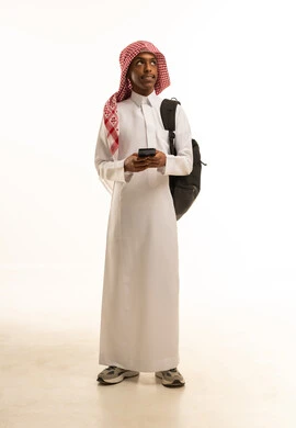 استخدام الأجهزة والتقنيات الحديثة والمتطورة, البحث والمطالعة من خلال الجوال، تصفح التطبيقات ووسائل التواصل الاجتماعي، بورتريه لشاب عربي خليجي سعودي مبتسم يرتدي الثوب التقليدي و الشماغ و حقيبة ظهر يستخدم الهاتف المحمول, خلفية بيضاء