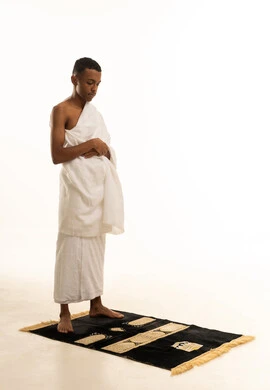 بورتريه من الجانب لشاب عربي خليجي سعودى يرتدي ثياب الإحرام يقف باستقامة على سجادة الصلاة بإيماءات الخشوع والتوسل والتضرع، أداء الصلوات على وقتها، التقرب الى الله والاجتهاد في العبادة، مناسك الحج والعمرة، الجسم بالطول الكامل، خلفية بيضاء