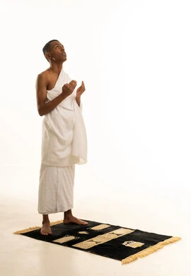 A portrait of a young Gulf Arab Saudi man wearing Ihram clothing, standing upright, raising his hands to the sky, praying with gestures of supplication and humility, embodying the concepts of the pillars of Islam and religious obligations, performing the rituals of Hajj and Umrah, full-body, white background.