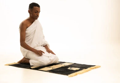 أداء الصلوات على وقتها، التقرب الى الله والاجتهاد في العبادة، مناسك الحج والعمرة، بورتريه من الجانب لشاب عربي خليجي سعودى يرتدي ثياب الإحرام يصلى على سجادة الصلاة بإيماءات الخشوع والتوسل والتضرع، خلفية بيضاء
