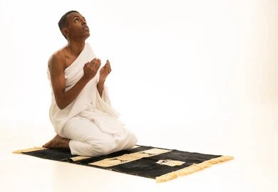 Drawing closer to God and praying, feeling tranquility and comfort, a portrait of a young Saudi Arabian Gulf man wearing Ihram clothing sitting on a prayer rug raising his hands upwards in humility, performing the rituals of Hajj and Umrah, white background.