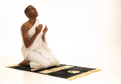 الدعاء والتقرب إلى الله، الشعور بالسكينة والراحة، بورتريه لشاب عربي خليجي سعودى يرتدي ثياب الإحرام يجلس على سجادة الصلاة يرفع يديه للأعلى يدعو بتضرع وخشوع، أداء مناسك الحج والعمرة، خلفية بيضاء